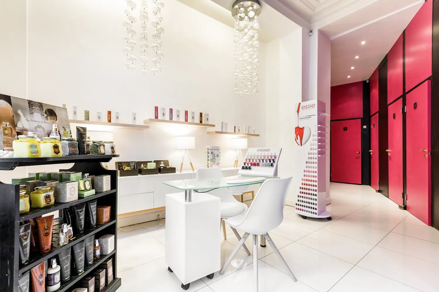 L'accueil de l'institut: sol blanc brillant, mur d'étagères de produits, table en verre et suspensions de perles, avec les portes roses des cabines au fond